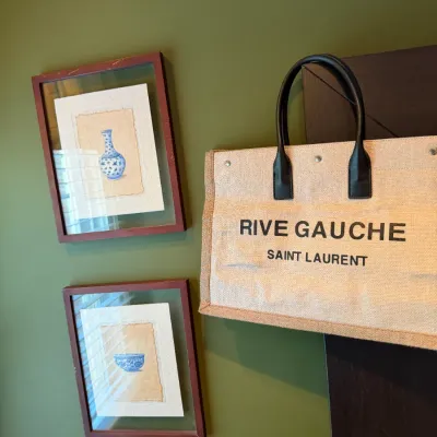 Saint Laurent Rive Gauche linen tote bag displayed alongside framed Chinese porcelain artwork on a green wall.