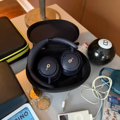 Sale Photo Thumbnail #105: Anker Soundcore over-ear wireless headphones resting in their black hardshell carrying case, displayed alongside a white Apple iPod Nano and other small electronics.