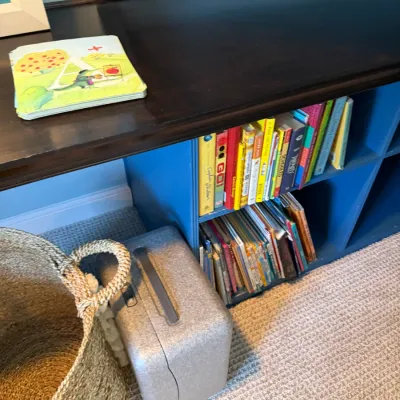 Low wooden bookcase painted blue with dark stained top, filled with children's books, shown alongside a woven storage basket and a...
