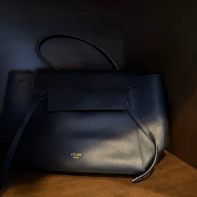 Black textured leather Céline Belt Bag featuring gold-tone logo lettering, a flap closure, top handle, and long side ties.