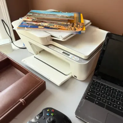 Sale Photo Thumbnail #106: White HP DeskJet all-in-one printer with a stack of postcards on top, sitting on a desk alongside a black Xbox controller and laptop.