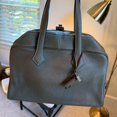 Taupe pebbled leather handbag featuring dual handles, contrast stitching, silver-tone hardware, and attached clochette with lock.