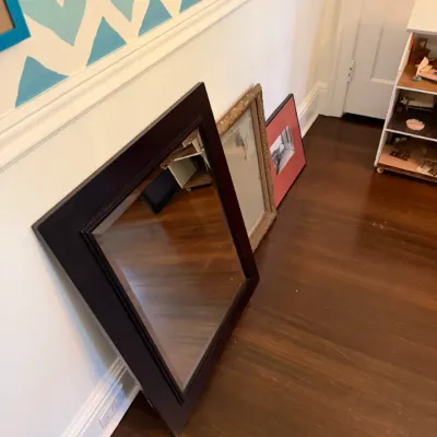 Collection of framed mirrors and wall art leaning against a wall, including a dark wood wall mirror and a ornate gilt-framed mirror.
