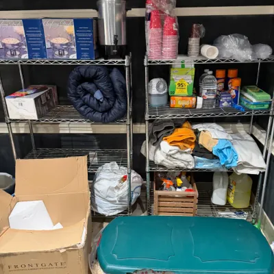 Sale Photo Thumbnail #263: Two metal wire utility shelving units stocked with various household goods, party supplies, cleaning products, and linens, alongside a large green plastic storage tote and a cardboard shipping box.