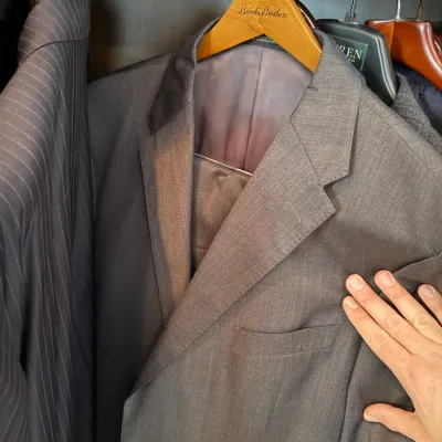 Sale Photo Thumbnail #242: Men's Brooks Brothers grey two-piece suit hanging on a wooden brand hanger alongside other suits.