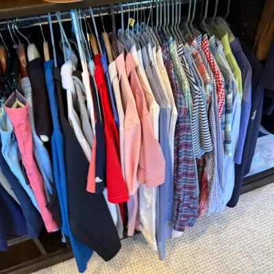 Closet rack filled with various men's button-down shirts, polo shirts, and jackets hanging below a shelf displaying handbags.
