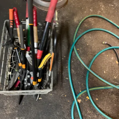 Sale Photo Thumbnail #217: Wire basket filled with various long-handled garden shears and loppers, shown alongside a coiled green garden hose on a concrete floor.