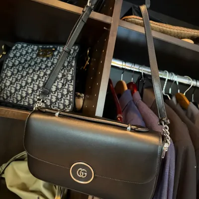 Black leather Gucci shoulder bag with silver-tone GG emblem, shown alongside other designer handbags in a closet arrangement.