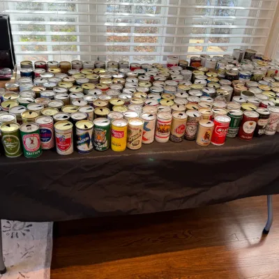 Sale Photo Thumbnail #153: Large collection of vintage pull-tab beer and ale cans displayed on a table, including brands like Rolling Rock, Carlsberg, Keith's, O'Keefe, and Schmidt's.