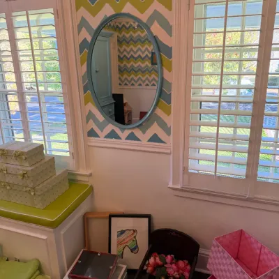 Oval wall mirror with a textured light blue frame, hung between two windows in a room with Chevron wallpaper.