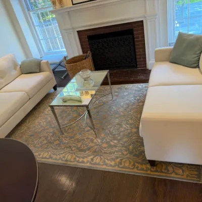 Sale Photo Thumbnail #130: Living room setup featuring two off-white upholstered sofas, a mirrored rectangular coffee table, and a large patterned area rug centered in front of a fireplace.