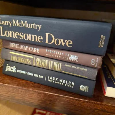 Sale Photo Thumbnail #257: Stack of four hardcover books without dust jackets resting on a wooden shelf, including titles such as Lonesome Dove by Larry McMurtry.