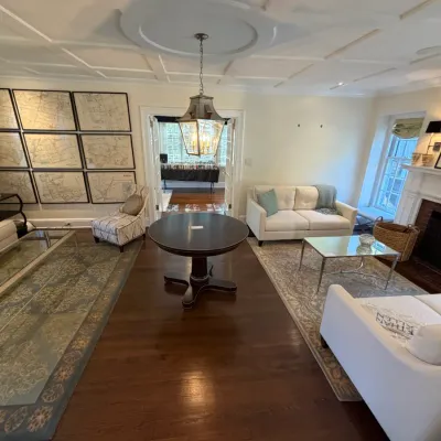 Spacious traditional living room featuring white upholstered sofas, a dark wood pedestal dining table, glass coffee tables, and a framed...