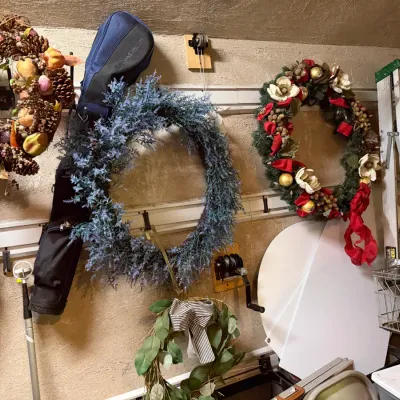 Sale Photo Thumbnail #220: Collection of decorative artificial wreaths including pinecone, purple flora, Christmas, and eucalyptus designs, hanging alongside a golf bag and step ladder.