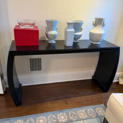 Sale Photo Thumbnail #123: Black minimalist console table with curved waterfall ends, topped with modern white and light blue ceramic vases and a clear glass bowl on a red box.