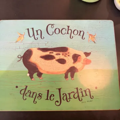 Sale Photo Thumbnail #199: French country style wooden placemat featuring a spotted pig illustration with the text 'Un Cochon dans le Jardin' by artist Julie Huber.