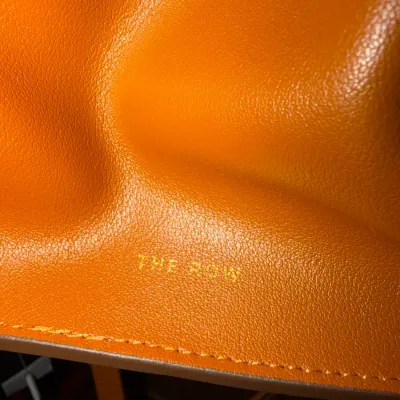 Close-up detail of a bright orange pebbled leather bag by The Row, featuring gold foil logo heat stamping near the stitched bottom edge.