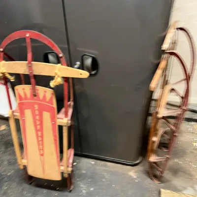Sale Photo Thumbnail #233: Pair of vintage wooden and metal snow sleds, including a Royal Racer model with red runners and steering bar, leaning against a gray storage cabinet.