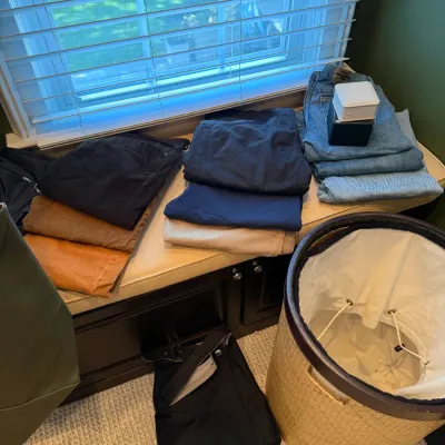 Selection of men's clothing including stacks of jeans, trousers, and corduroy pants displayed on a padded window bench alongside a...