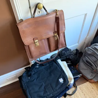 Vintage brown leather briefcase hanging on a door, shown above a pile of various nylon duffel bags and backpacks.