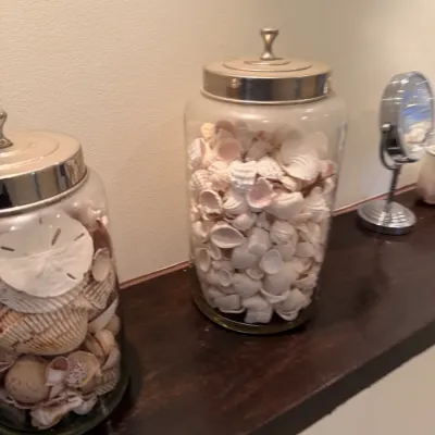 Two large glass apothecary jars filled with sorted sea shells, featuring brushed metal lids with finials, displayed alongside a small...