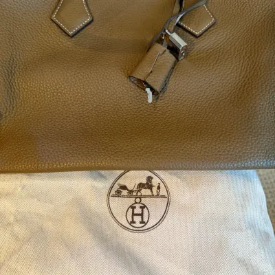Hermès taupe pebbled leather handbag with contrast stitching, silver-tone lock and key clochette, and original branded dust bag.