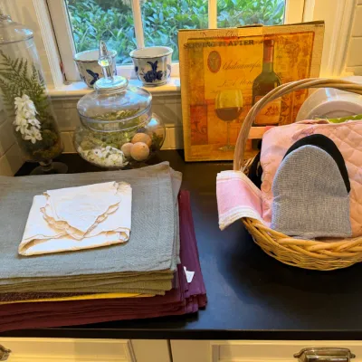Sale Photo Thumbnail #177: Kitchen table linens, including stacked placemats, vintage glass terrarium jars, a woven wicker basket filled with oven mitts, and decorative home accessories on a countertop.