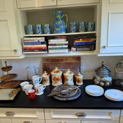 Sale Photo Thumbnail #179: Kitchen counter and shelving display featuring floral ceramic canisters, pitchers, cookbooks, serving plates, mugs, and a wooden tiered stand.