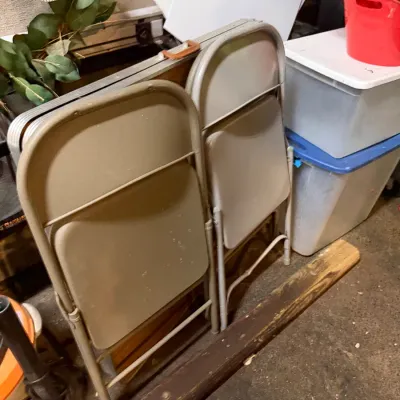 Sale Photo Thumbnail #219: Pair of tan metal folding chairs standing upright in storage next to a folded banquet table and plastic storage totes.