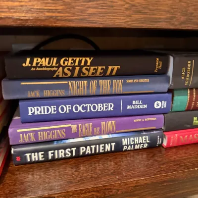 Sale Photo Thumbnail #254: Stack of hardcover books on a wooden shelf, including titles by J. Paul Getty, Jack Higgins, and Michael Palmer.
