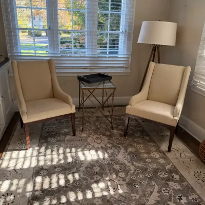 Pair of cream upholstered wingback accent chairs with dark wood legs, arranged on a patterned area rug alongside a glass-top side table...