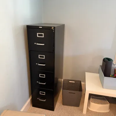 Tall four-drawer black metal filing cabinet with silver hardware, positioned in an office corner beside a plastic storage bin and small...