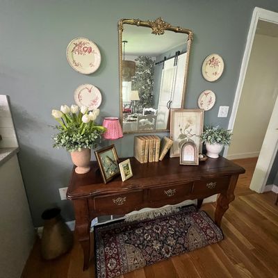 Sale Photo Thumbnail #4: French Country style dark wood console table with decorative wall plates, an ornate gilt mirror, set of books, and artificial floral displays.