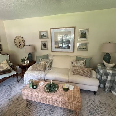 Sale Photo Thumbnail #2: Traditional living room setup featuring an off-white three-cushion sofa, a wicker coffee table, an armchair with wood trim, and various table lamps and framed wall art.