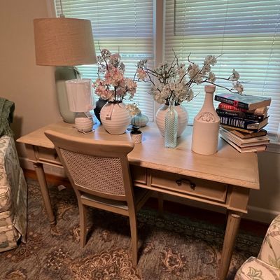 Sale Photo Thumbnail #7: Light-finished wooden writing desk paired with a cane-back dining or accent chair, decorated with table lamps, ceramic vases with faux floral arrangements, and stacked books.