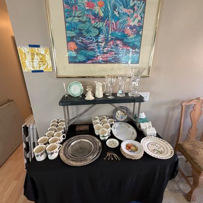 Display table featuring vintage teacups, silver-plated platters, floral china, crystal glassware, and brass keys, set beneath a large...