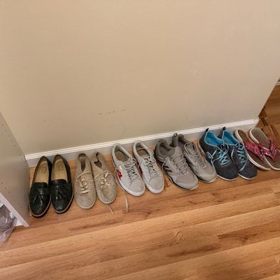 A lineup of women's shoes neatly arranged along a wall on wood floors, including loafers, lace-up sneakers, athletic shoes, and...