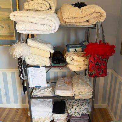 Rolling metal utility rack organized with stacked bed linens, blankets, comforters, and decorative bags.