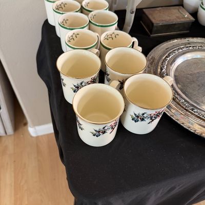 Collection of holiday-themed ceramic mugs with holly accents and gold rims displayed alongside silver-plated serving trays.