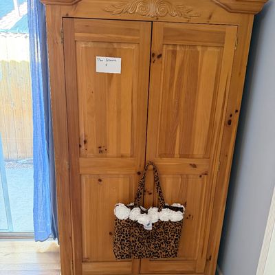 Large wooden pine armoire featuring an arched top with carved detailing, two paneled cabinet doors, and a leopard print tote bag hanging...
