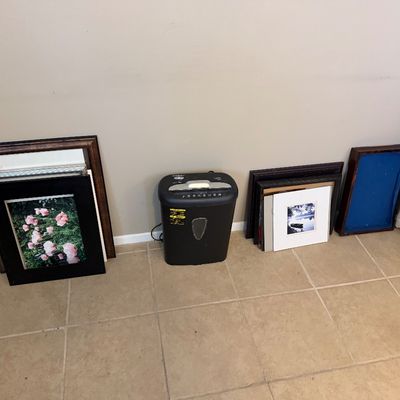 A black paper shredder positioned between several framed art prints and empty picture frames along a wall.