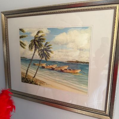 Framed tropical beach art print featuring palm trees and wooden boats anchored along a shoreline, presented in a matted metallic-tone frame.