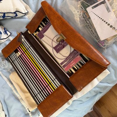 Brown faux-leather folding case containing an extensive knitting needle set with additional accessories, including circular needles,...