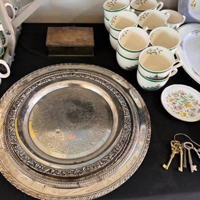 Collection of tabletop items including large ornate silver-plated trays, stacked green-rimmed china teacups, a small trinket dish, brass...