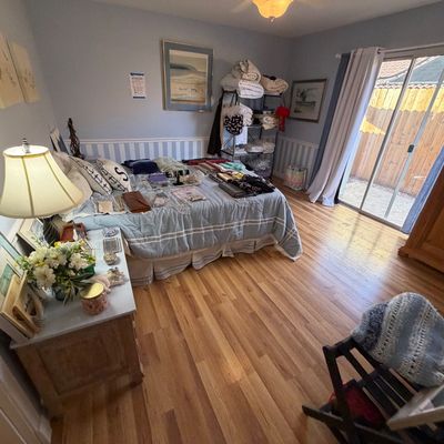 Bedroom featuring a double bed laid out with clothing and linens, accompanied by a wooden armoire, nightstand with lamp, and metal...