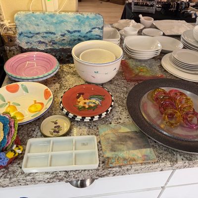 Collection of assorted kitchenware and decor, including ceramic bowls, plates, colorful napkin rings, and crocheted coasters.