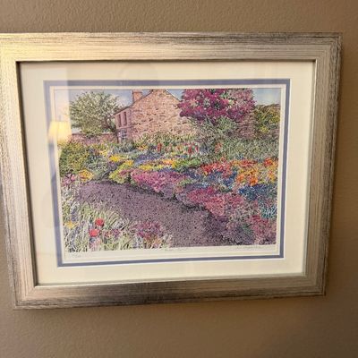 Framed and matted art print depicting a vibrant cottage garden and stone house, finished in a silver-toned frame.
