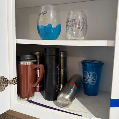 Assorted drinkware inside a kitchen cabinet, including printed stemless wine glasses, travel mugs, and stainless steel water bottles.