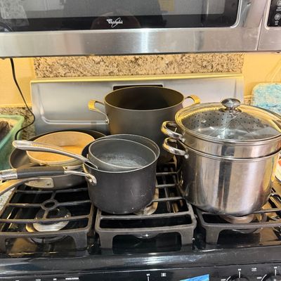 Assorted cookware resting on a black gas stovetop, including non-stick pots, a skillet, and a large stainless steel steamer pot with a...