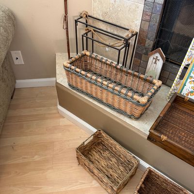 Assorted woven baskets and fireplace accessories arranged on a hearth, including a long rectangular rattan basket and a dark wood tray.
